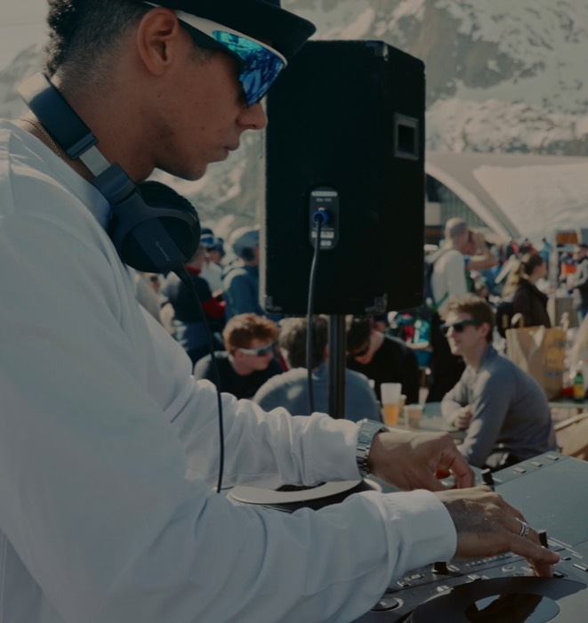 Un DJ con gafas de sol y auriculares pinchando al aire libre, con esquiadores y cumbres nevadas detrás