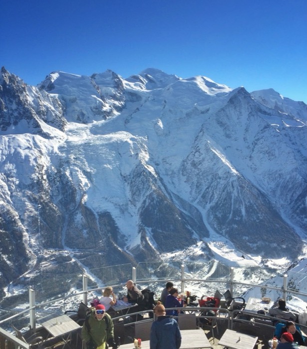 Personas en mesas de una terraza de montaña, con el macizo del Mont Blanc nevado detrás bajo un cielo azul intenso